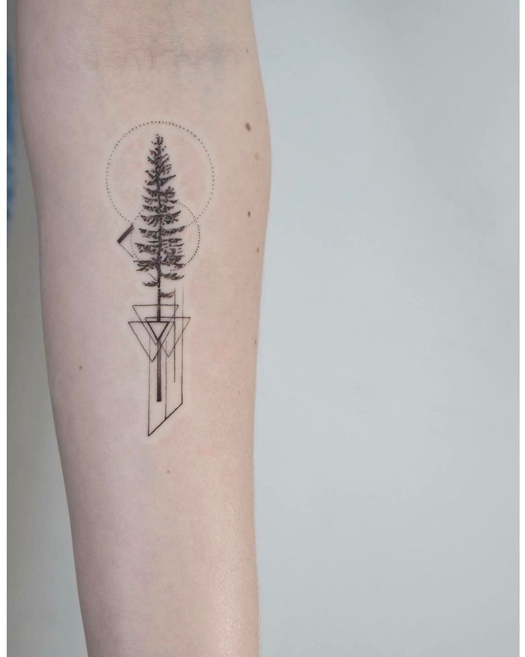 A minimalist black ink tattoo of a pine tree with geometric shapes on an arm.