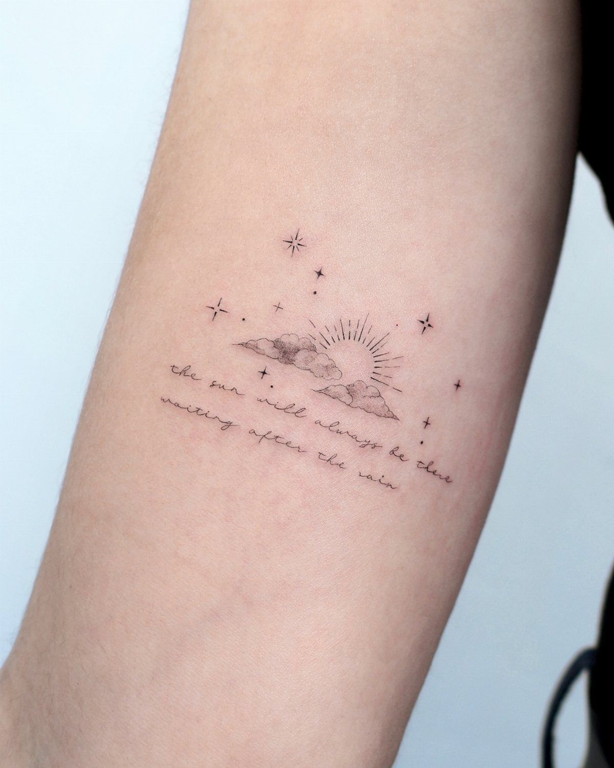 A fine line black ink tattoo of a sun, clouds, and stars with a cursive quote on a forearm.