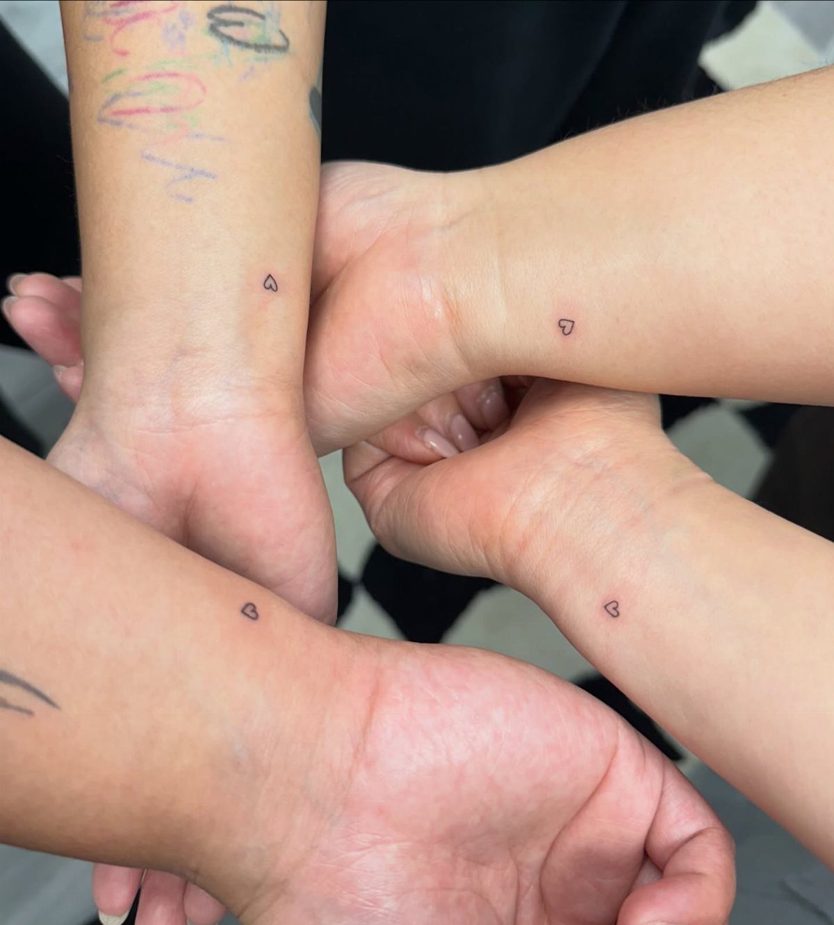 Four wrists joined in a circle, each displaying a tiny black heart tattoo.