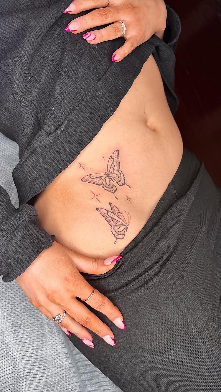 Two small fine-line butterfly tattoos on a person's hip with pink manicured nails.