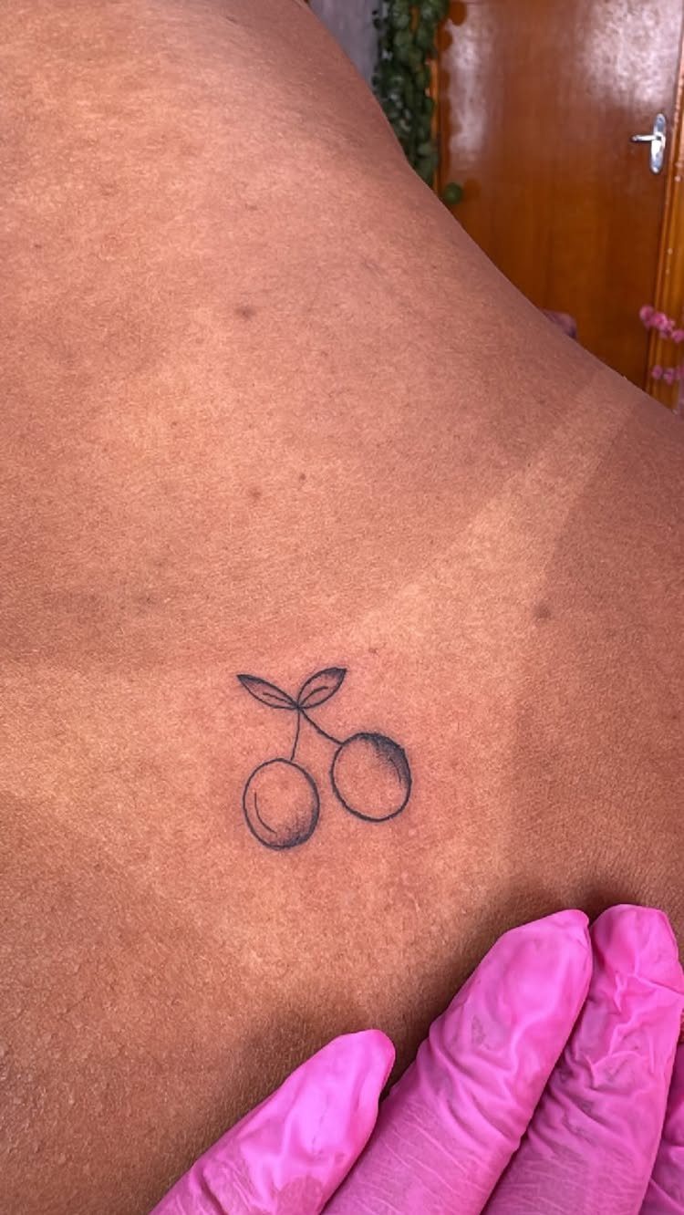A small black ink cherry tattoo on a person's skin being touched by a pink gloved hand.