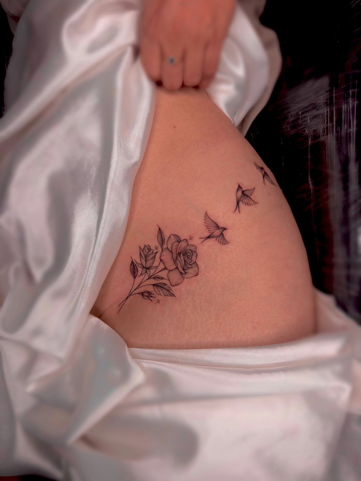 A black ink rose and bird tattoo on a person's hip with white satin fabric.
