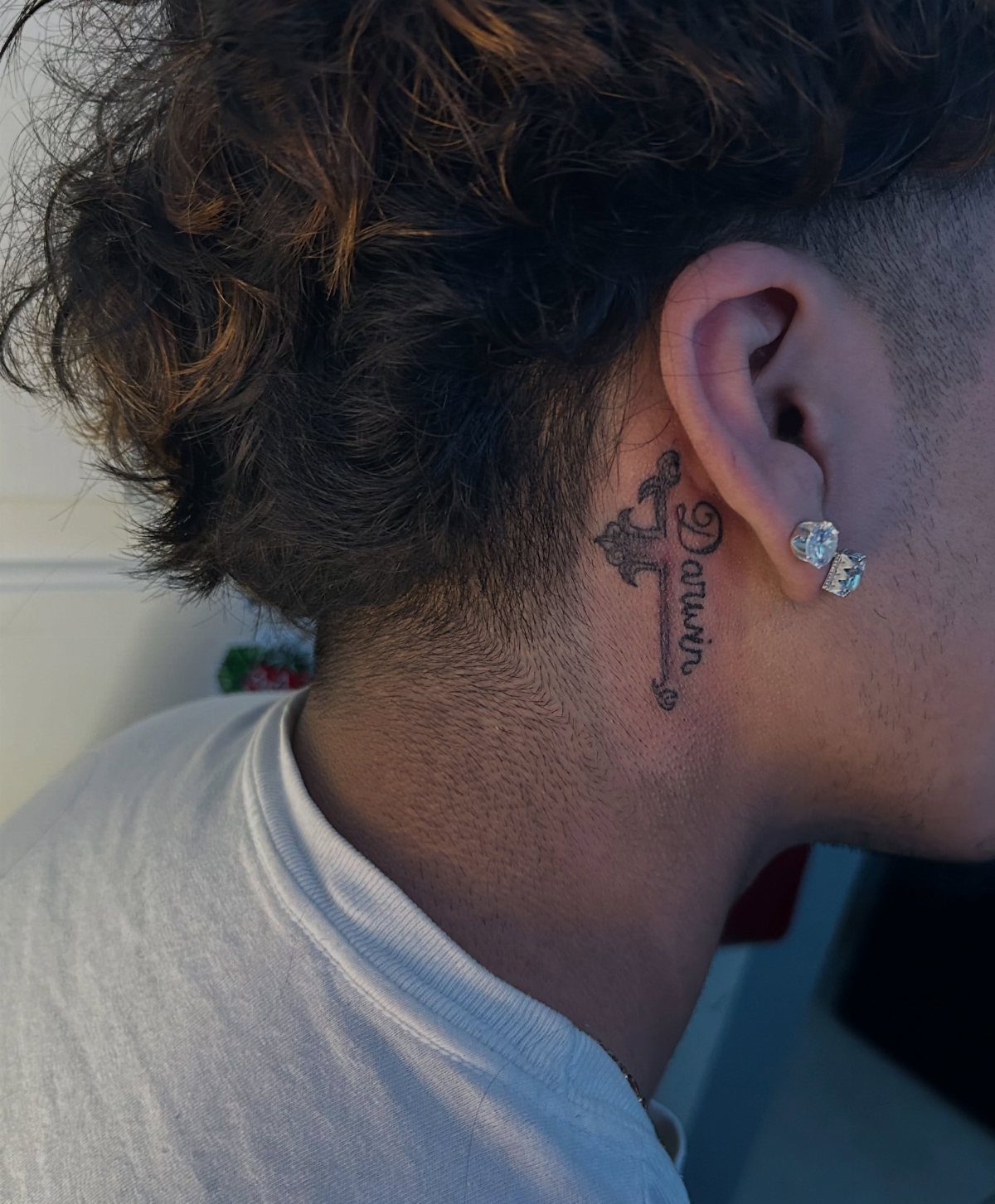 15 Small Tattoo Placement Ideas for Every Style 2 A small cross tattoo with the name Darwin placed behind a person's ear.