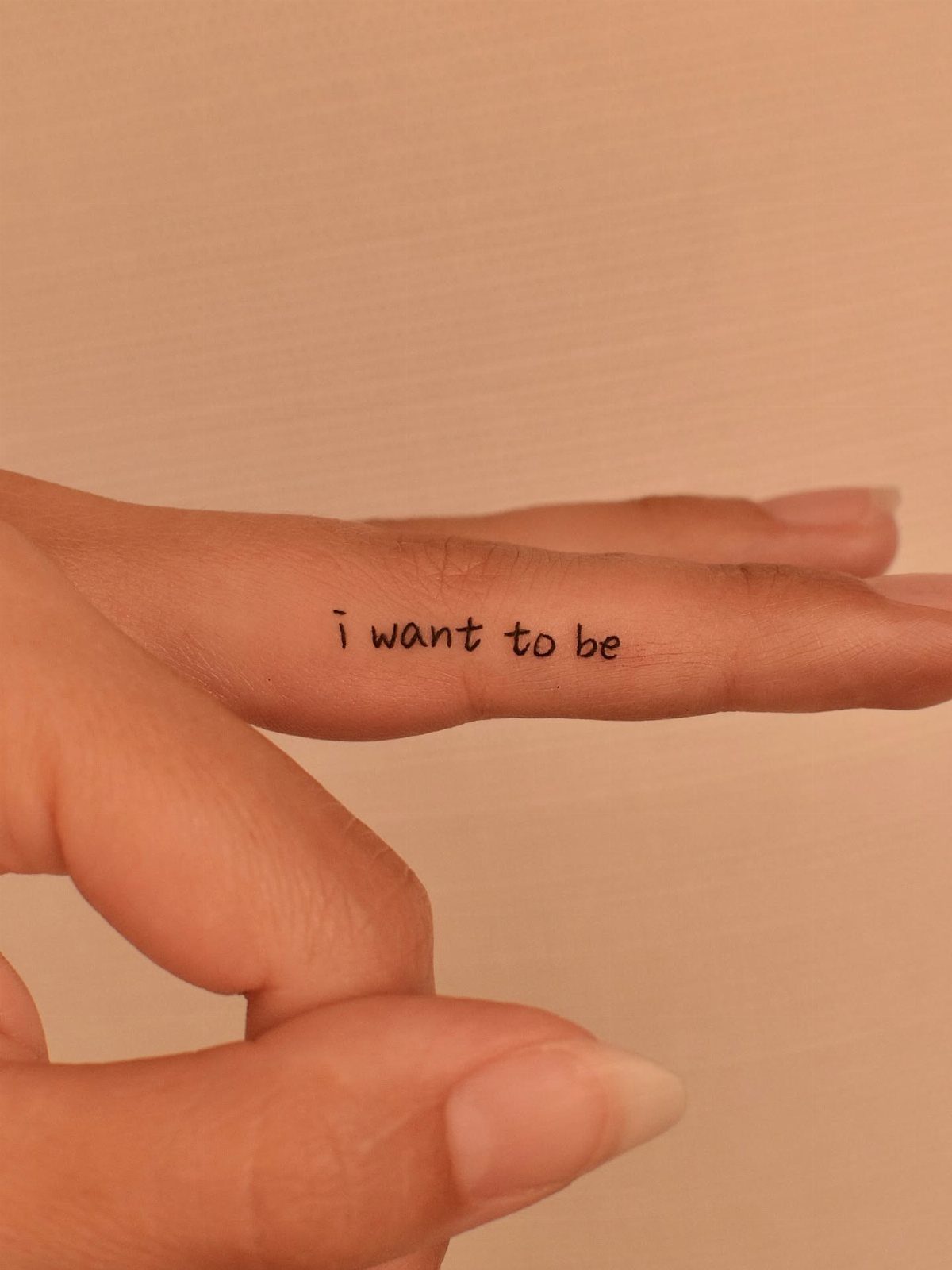 A small black ink tattoo reading 'i want to be' on the side of a person's finger.