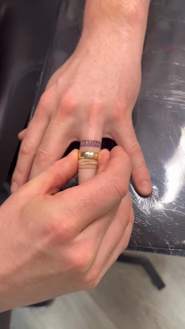 A close-up of a hand featuring a small Roman numeral tattoo on the ring finger.