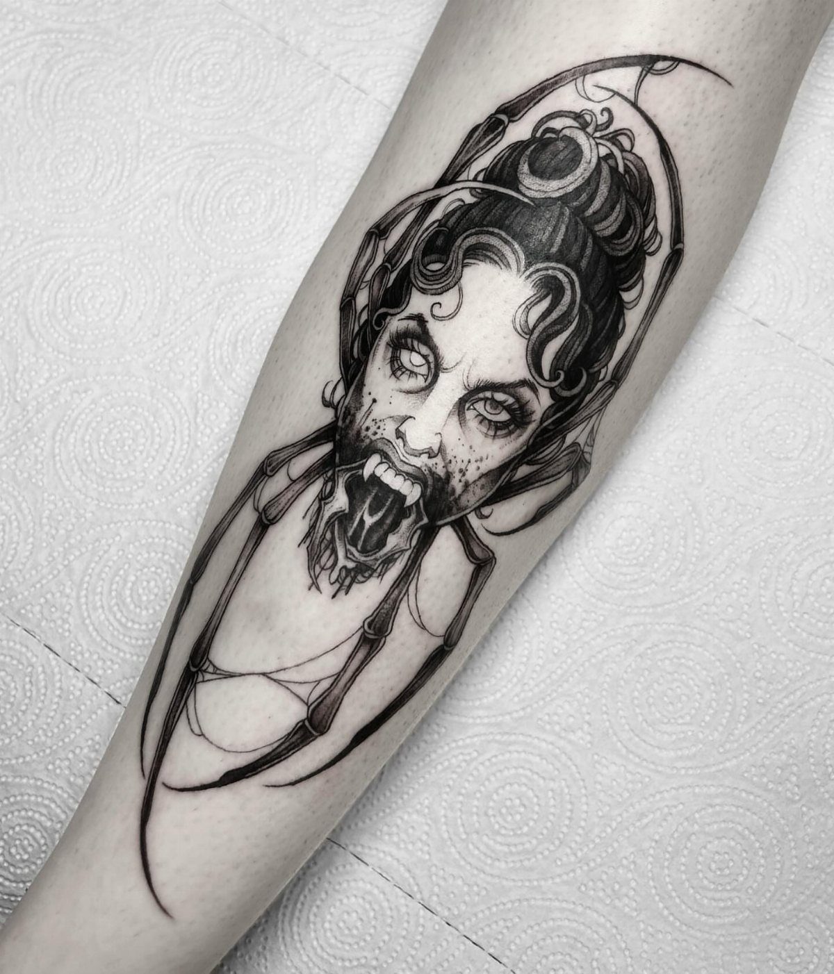 A black and grey tattoo of a woman's face combined with a spider on a leg.