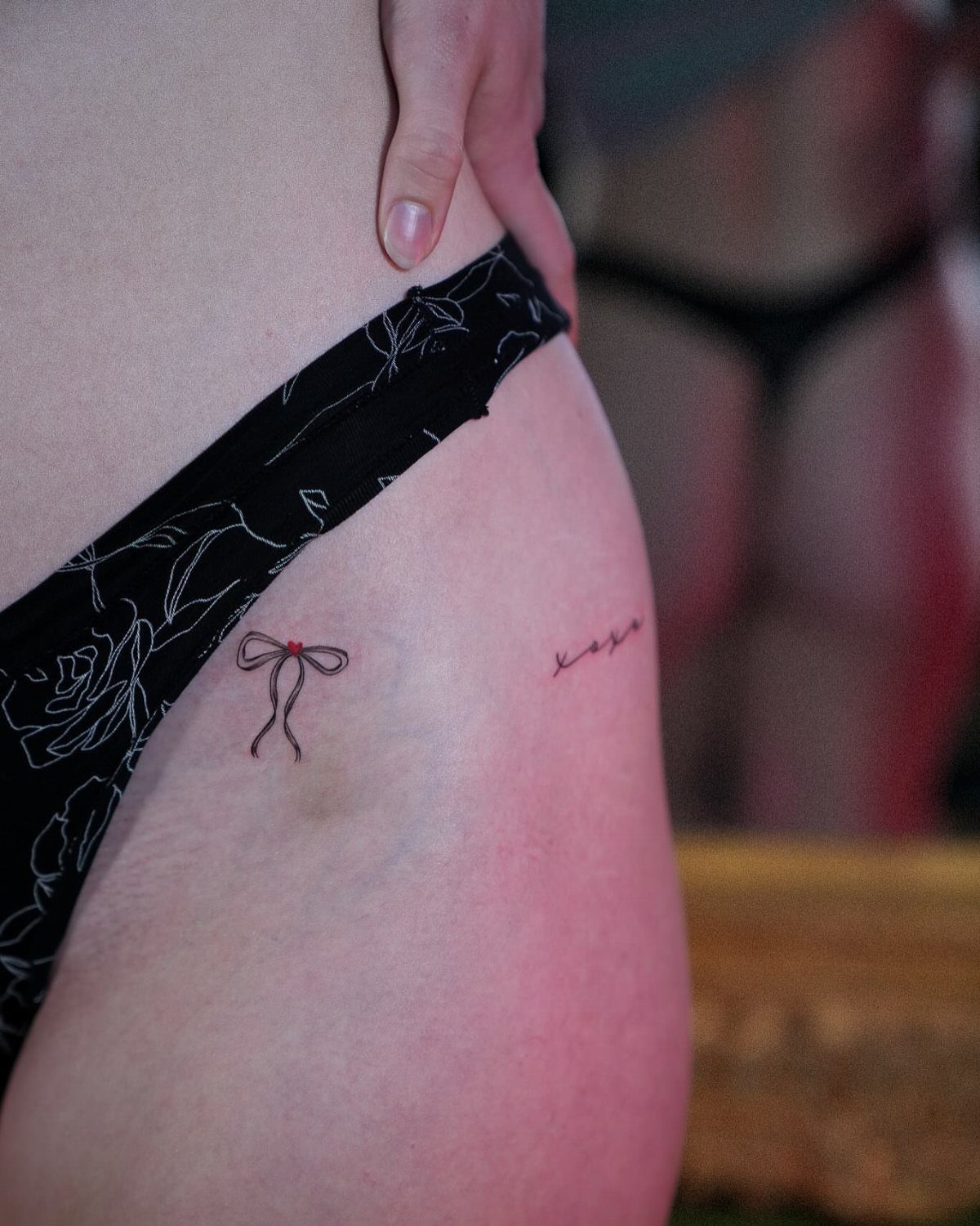 Small fine-line tattoos of a bow and script on a person's hip and pelvic area.