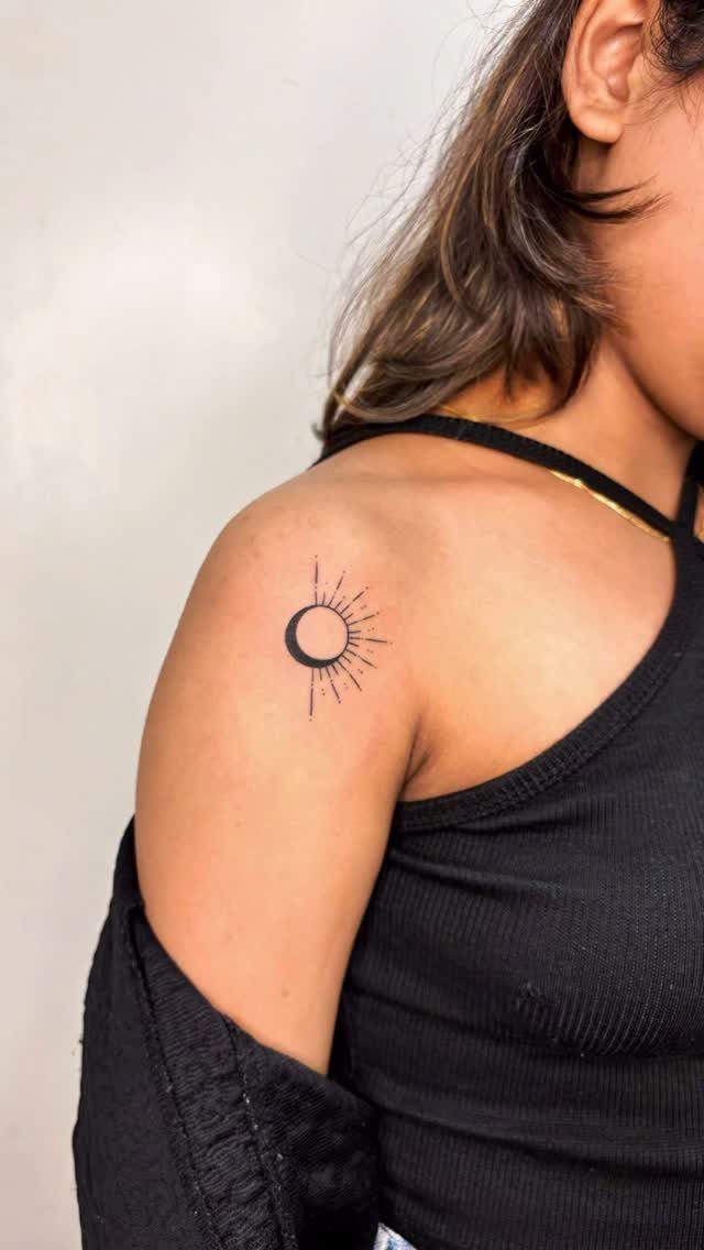 A small crescent moon and sunbeam tattoo on a person's shoulder.
