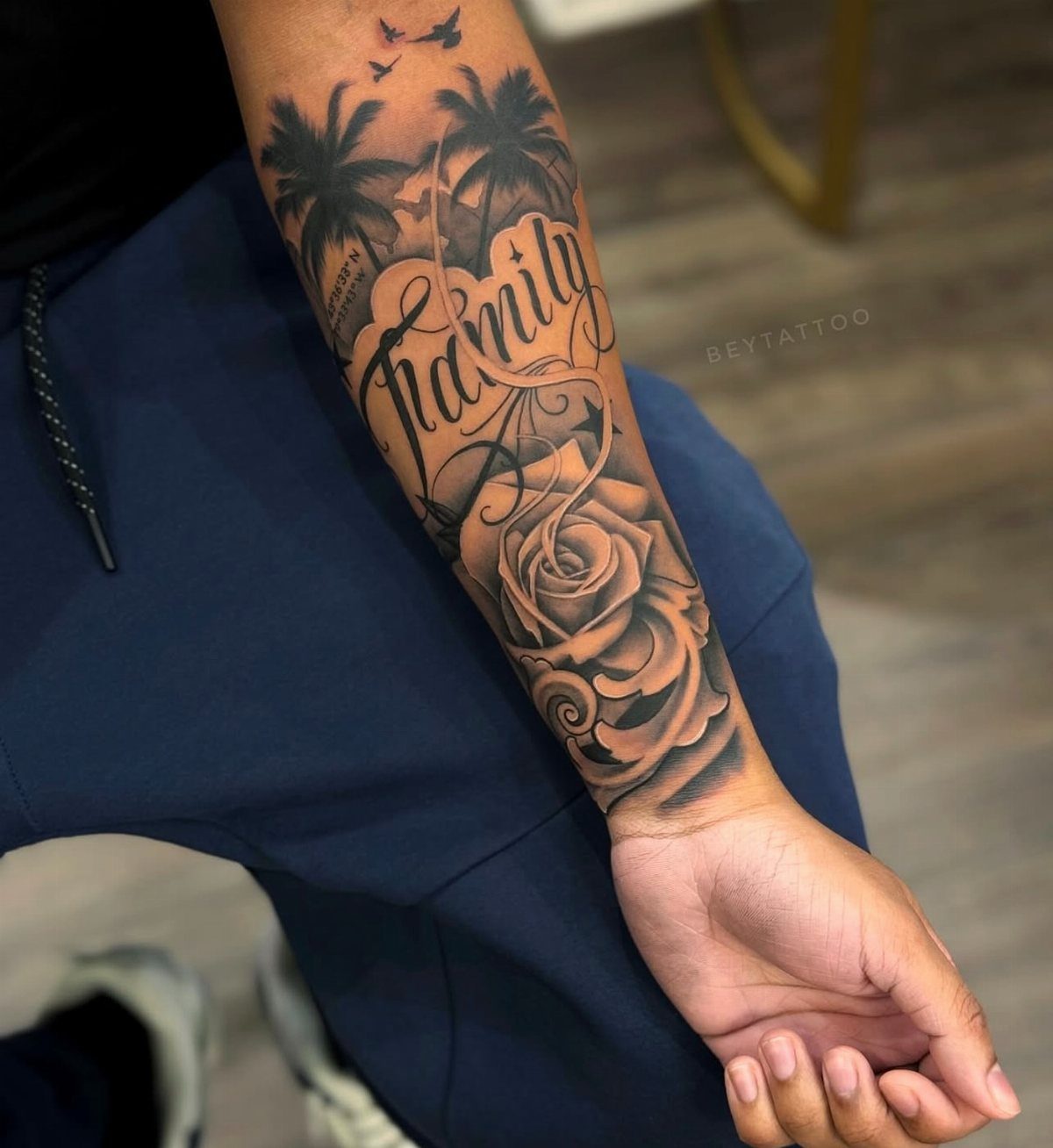 A detailed black and grey forearm tattoo featuring script text, palm trees, and a rose.