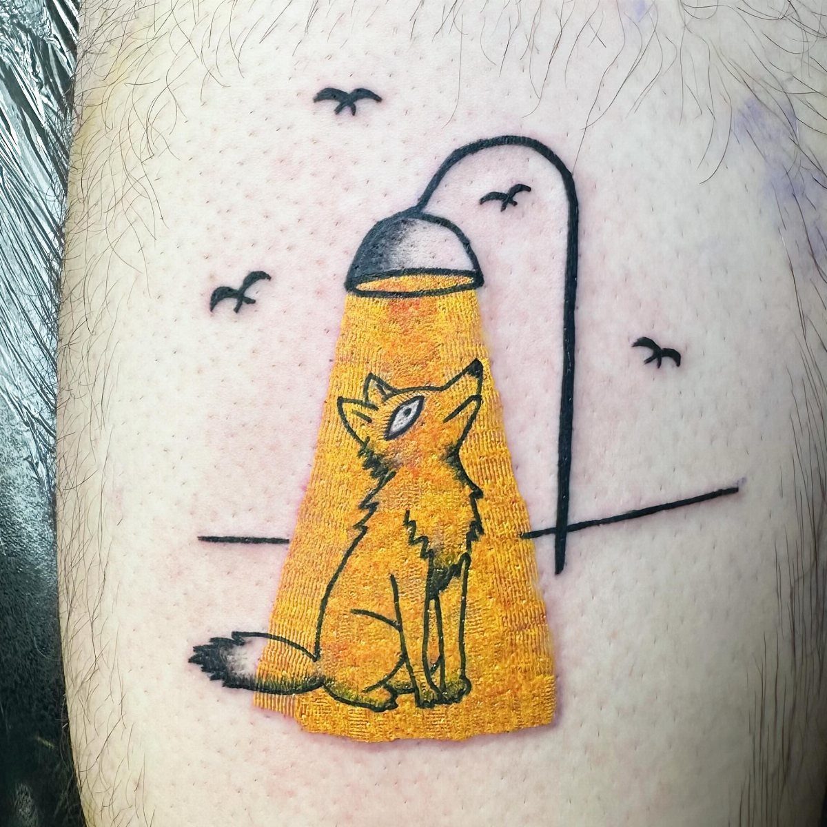 A tattoo of a yellow fox sitting under a streetlamp with birds flying in the background.