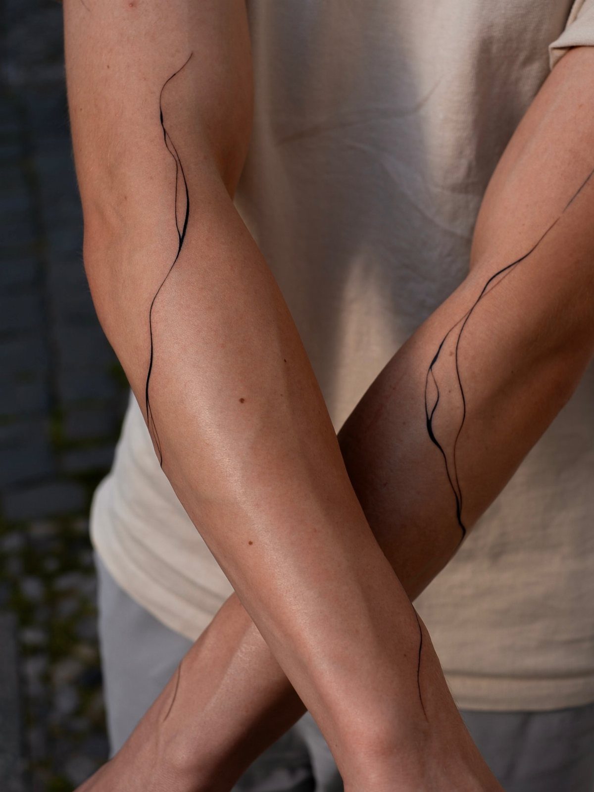 A person's crossed arms featuring minimalist black wavy line art tattoos on the forearms.