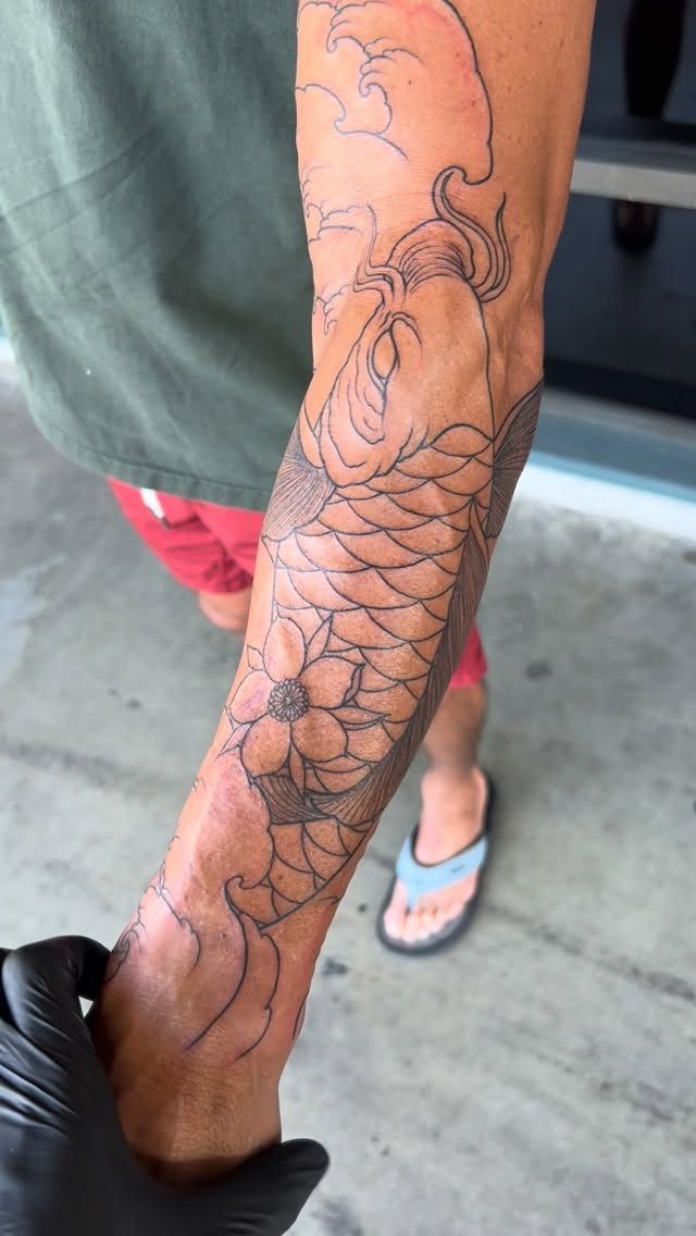 14 Tattoo Style Outline Ideas for Your Next Appointment 10 A large black ink koi fish and flower tattoo outline on a person's forearm.