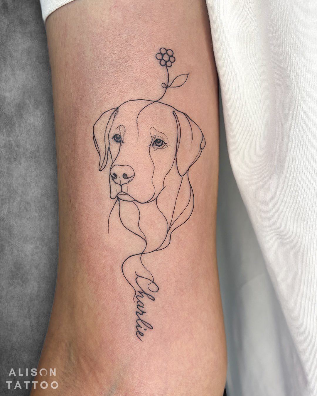 14 Tattoo Style Outline Ideas for Your Next Appointment 5 A minimalist fine line tattoo of a dog's face with a flower and the name Charlie.
