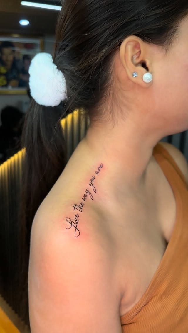 A fine line script tattoo reading 'Live the way you are' on a person's shoulder.