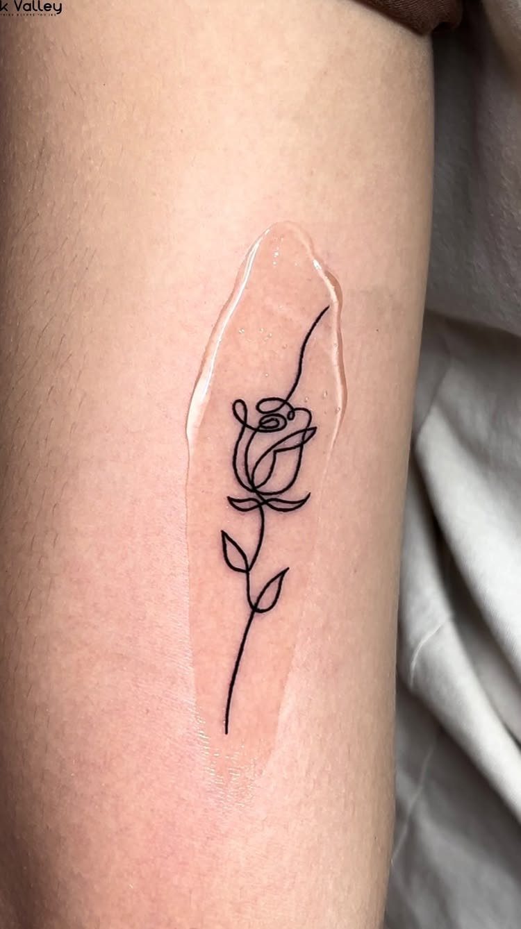 A black ink continuous line tattoo of a rose on human skin.