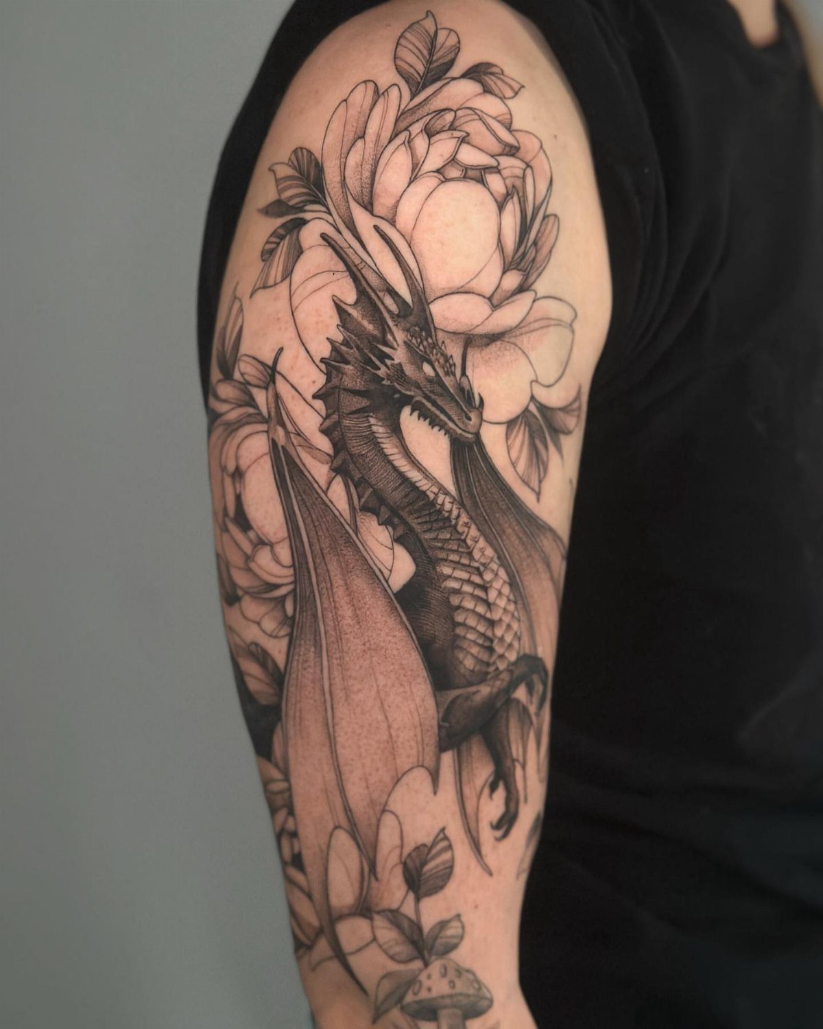 A detailed black and grey dragon tattoo with large wings and peony flowers on an arm.