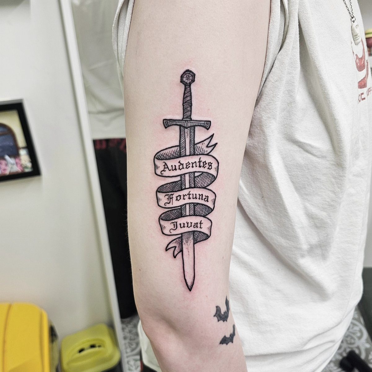 A black ink woodcut style tattoo of a sword with a banner on an arm.