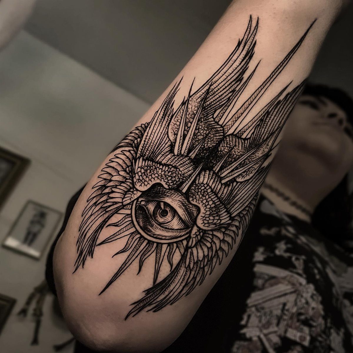 A black ink woodcut style tattoo of a winged eye on a person's arm.