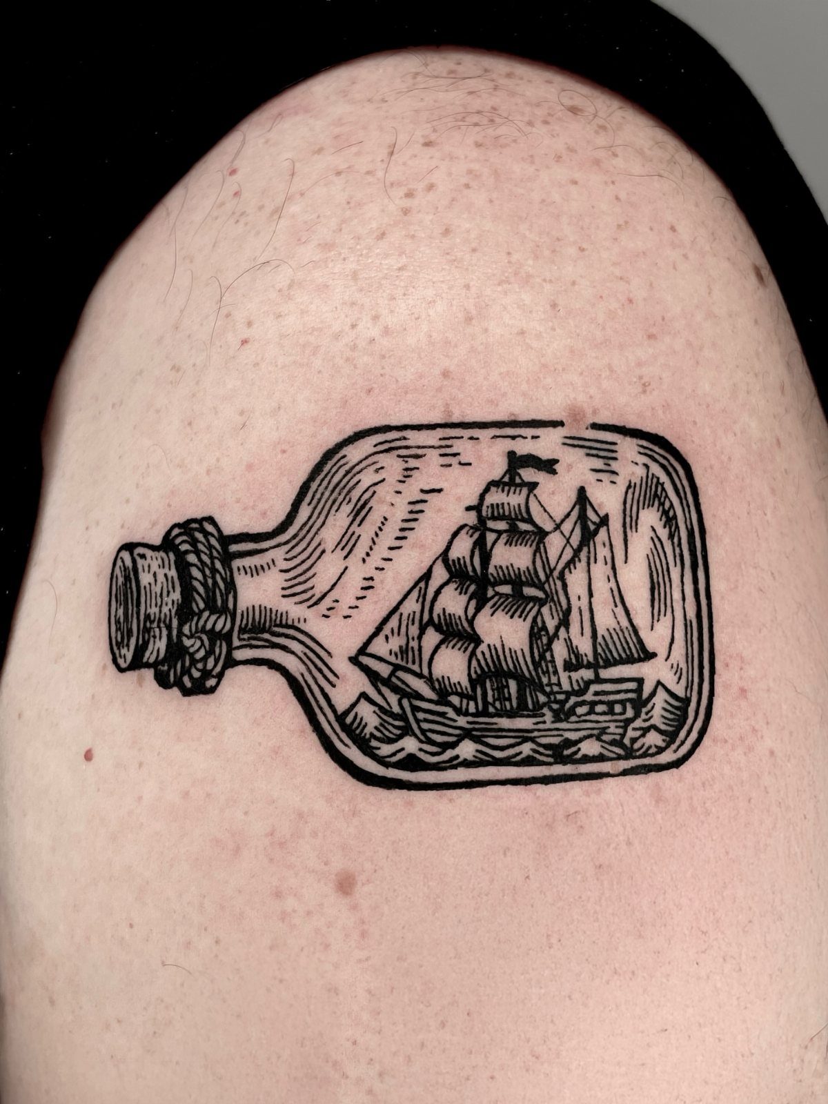 A woodcut style tattoo of a sailing ship contained within a glass bottle on skin.