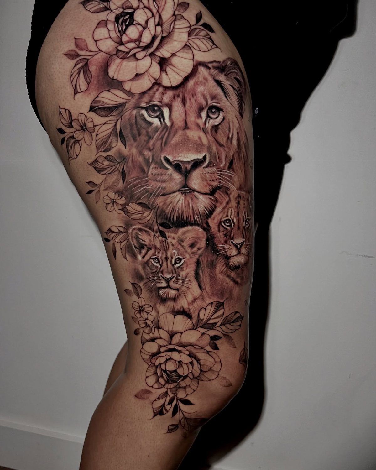 A large thigh tattoo depicting a lion, two cubs, and blooming peony flowers in black and grey.