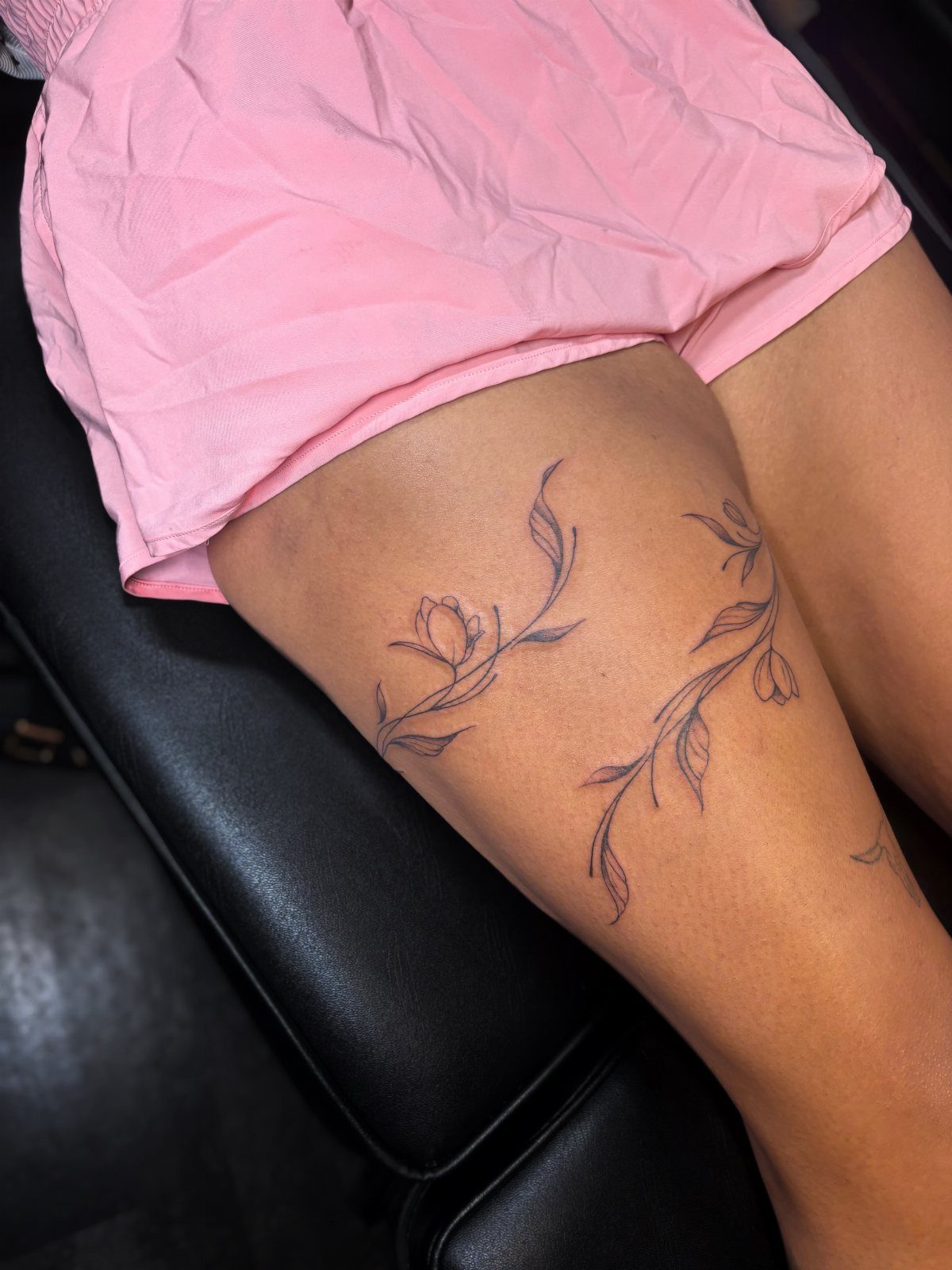 Fine line black ink vine tattoos with small leaves and flowers on a person's thigh.