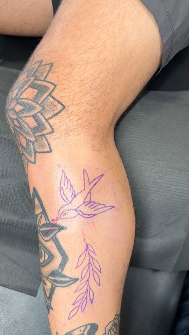 A person's leg featuring existing black tattoos and a new purple ink bird stencil.