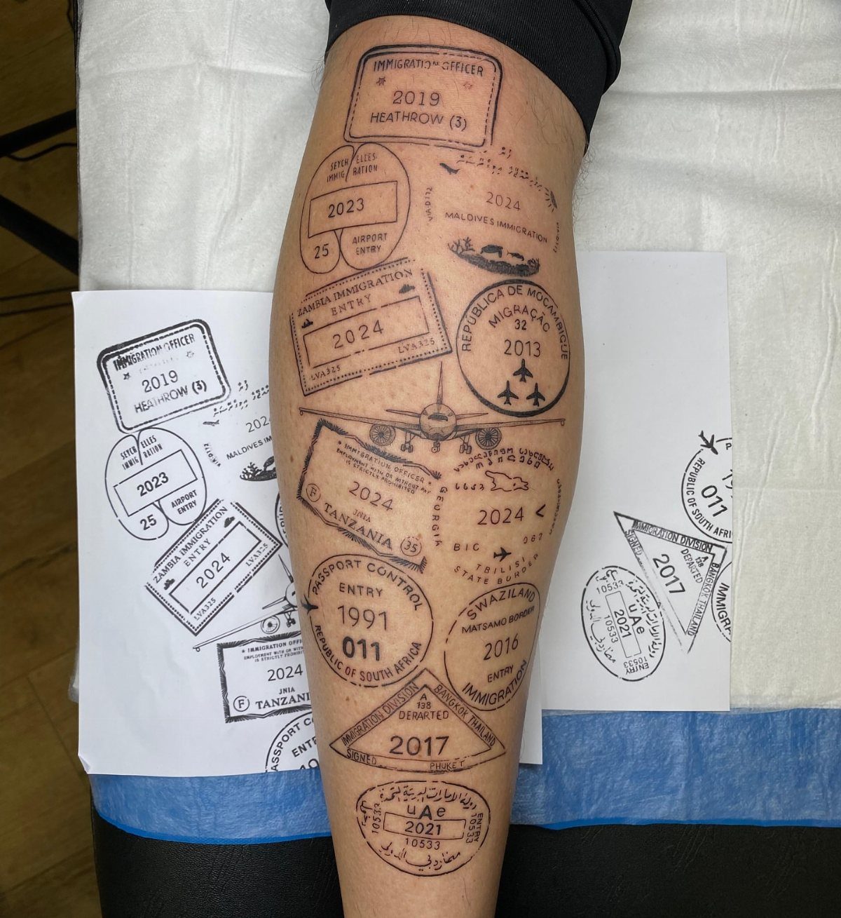 A person's leg featuring several black ink tattoos designed as various international passport entry stamps.