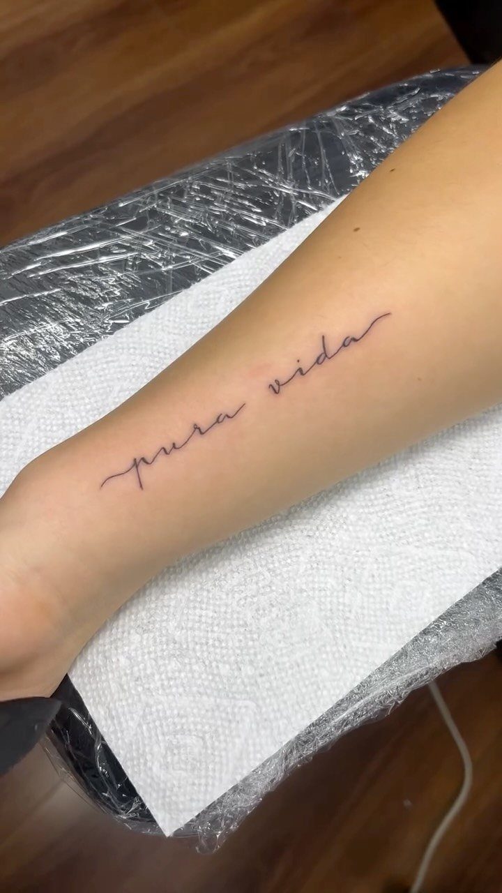 A forearm with a fine-line script tattoo reading pura vida in black ink.