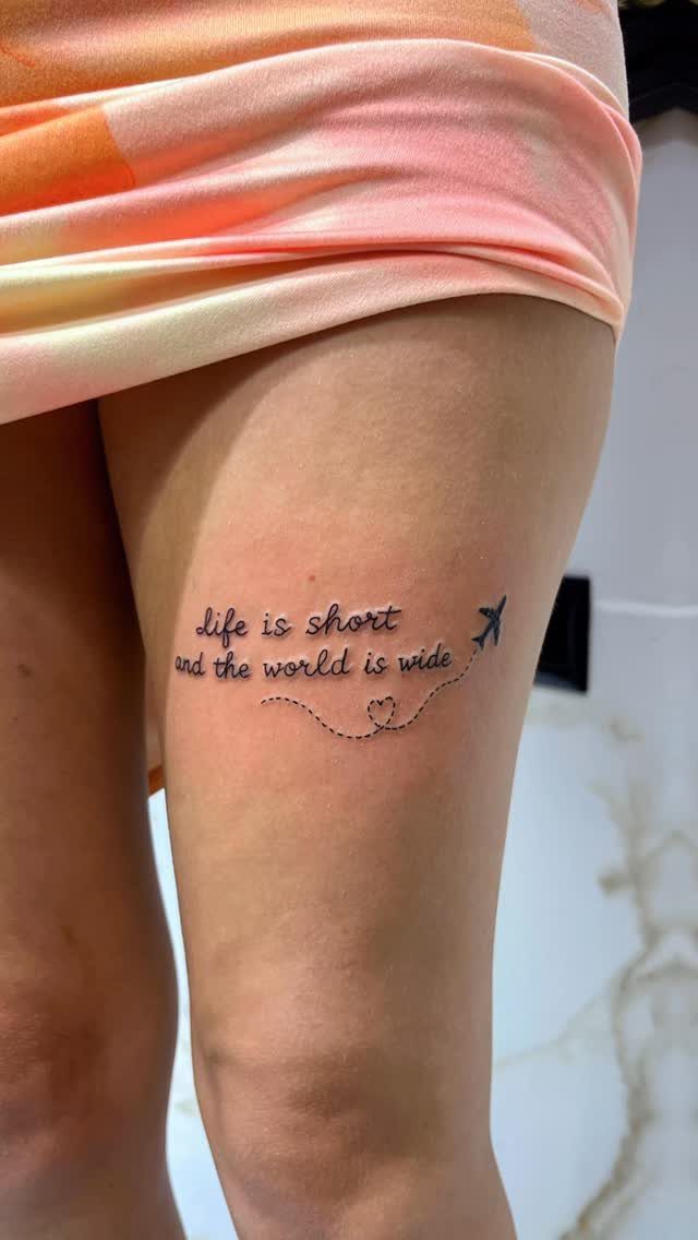 A black ink tattoo on a person's thigh reading life is short and the world is wide.