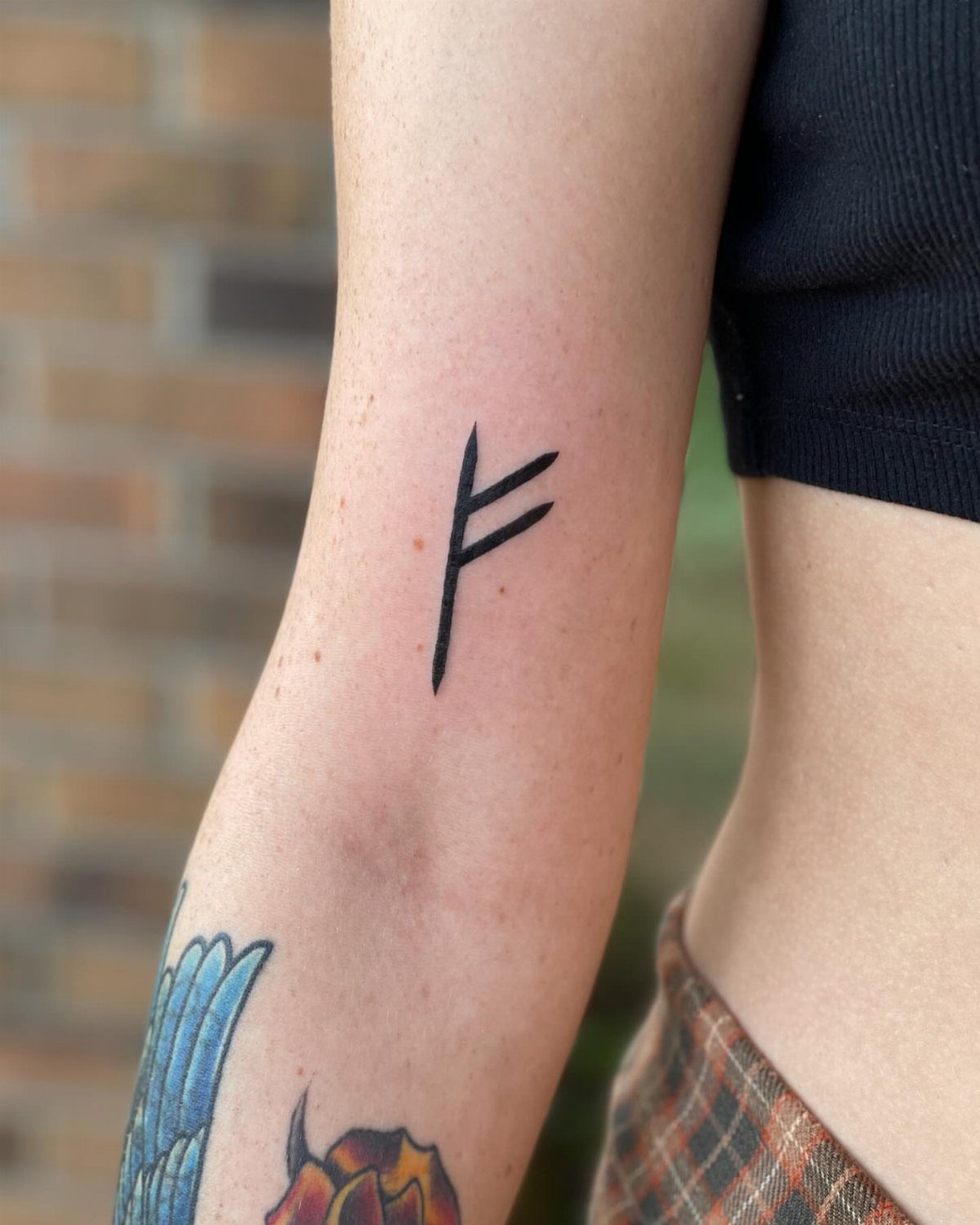 A black ink Fehu rune tattoo on a person's forearm against a brick background.