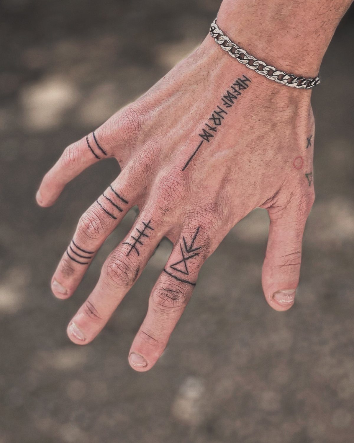 A hand with black ink runic tattoos and a silver chain bracelet on the wrist.