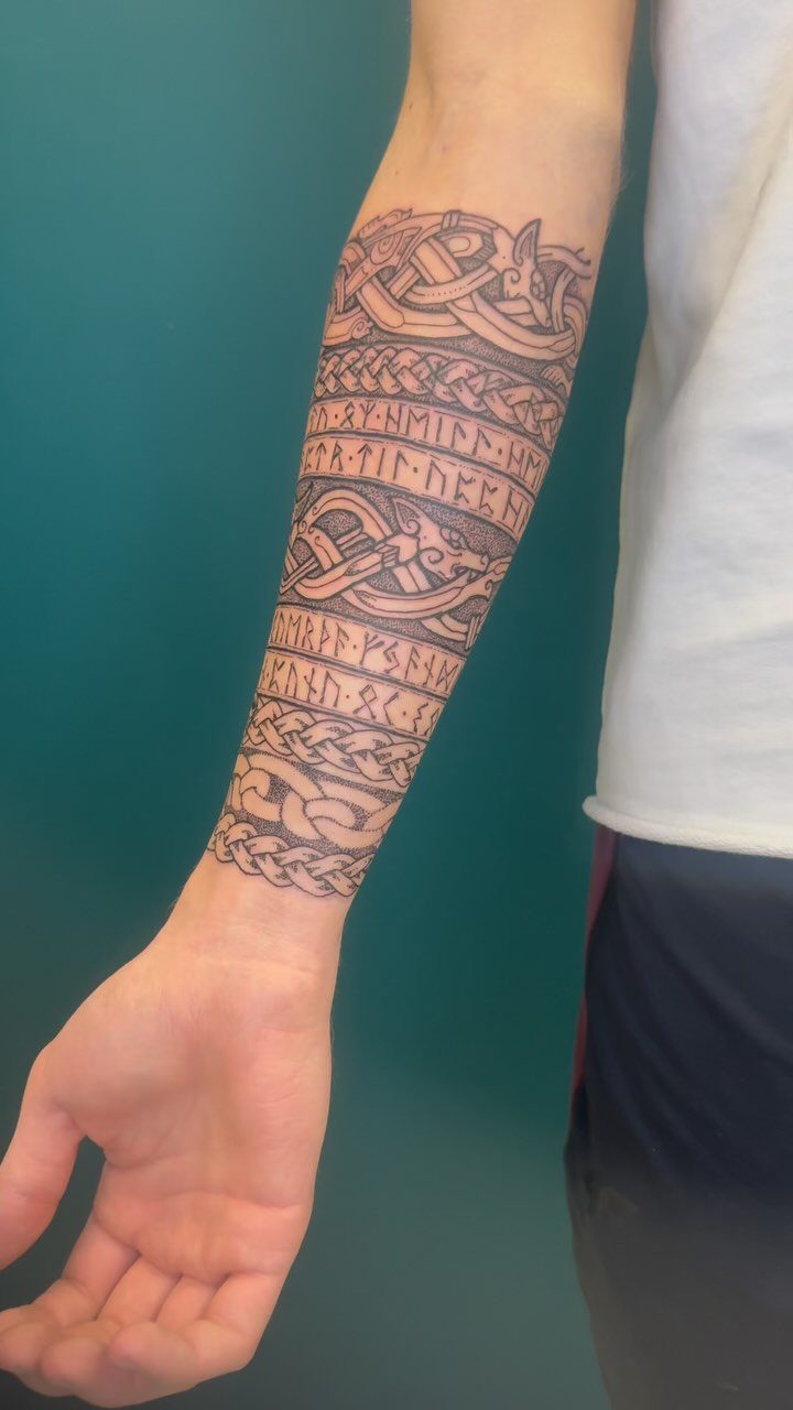 A forearm tattoo featuring intricate knotwork, animal motifs, and rows of Viking runes in black ink.