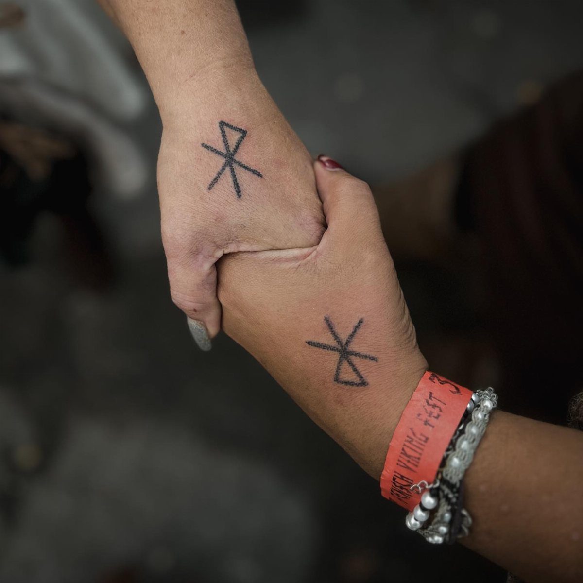 Two hands holding each other, displaying black Viking rune tattoos on their wrists.