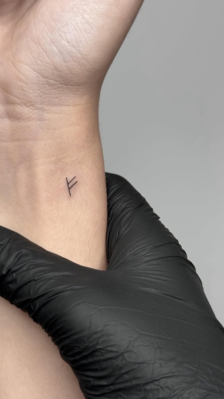 A small black Viking rune tattoo on a person's wrist held by a gloved hand.