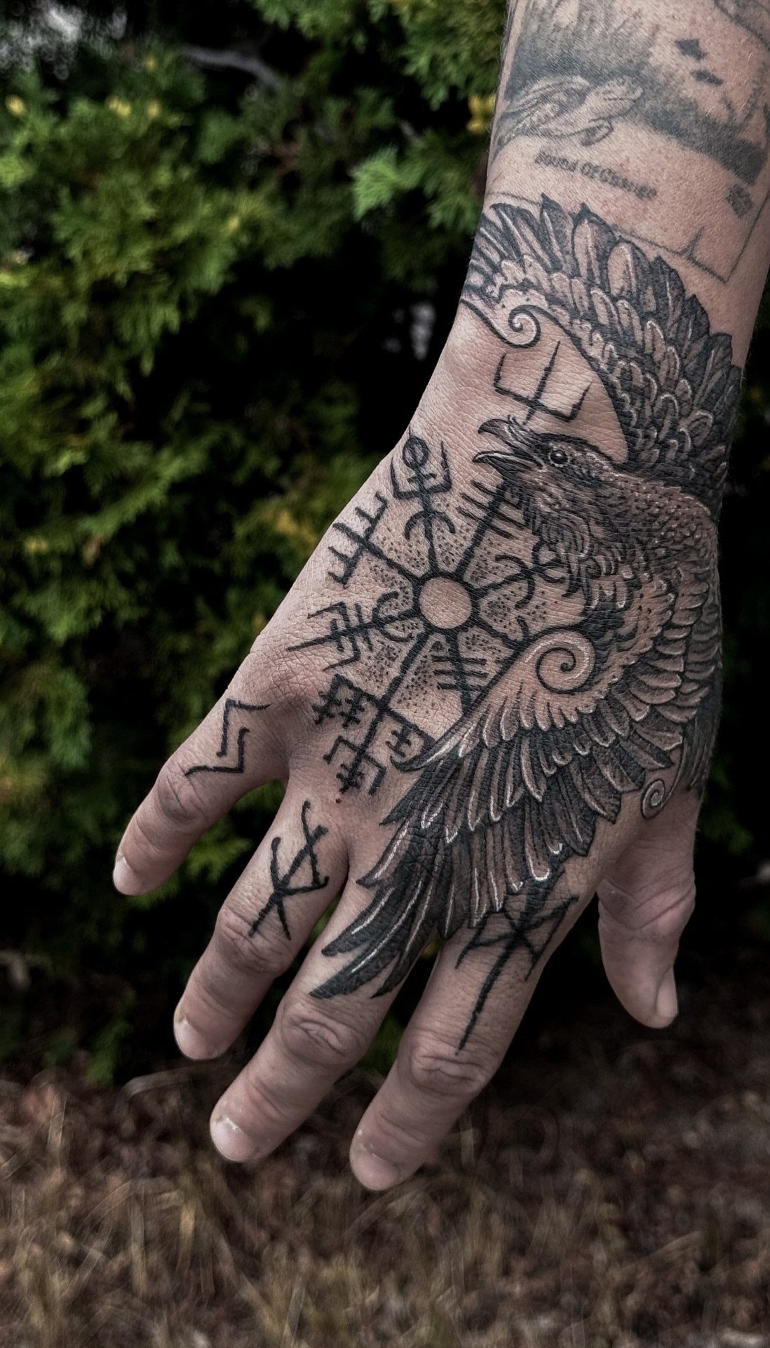 A hand and forearm decorated with a large raven tattoo and various Viking rune symbols.