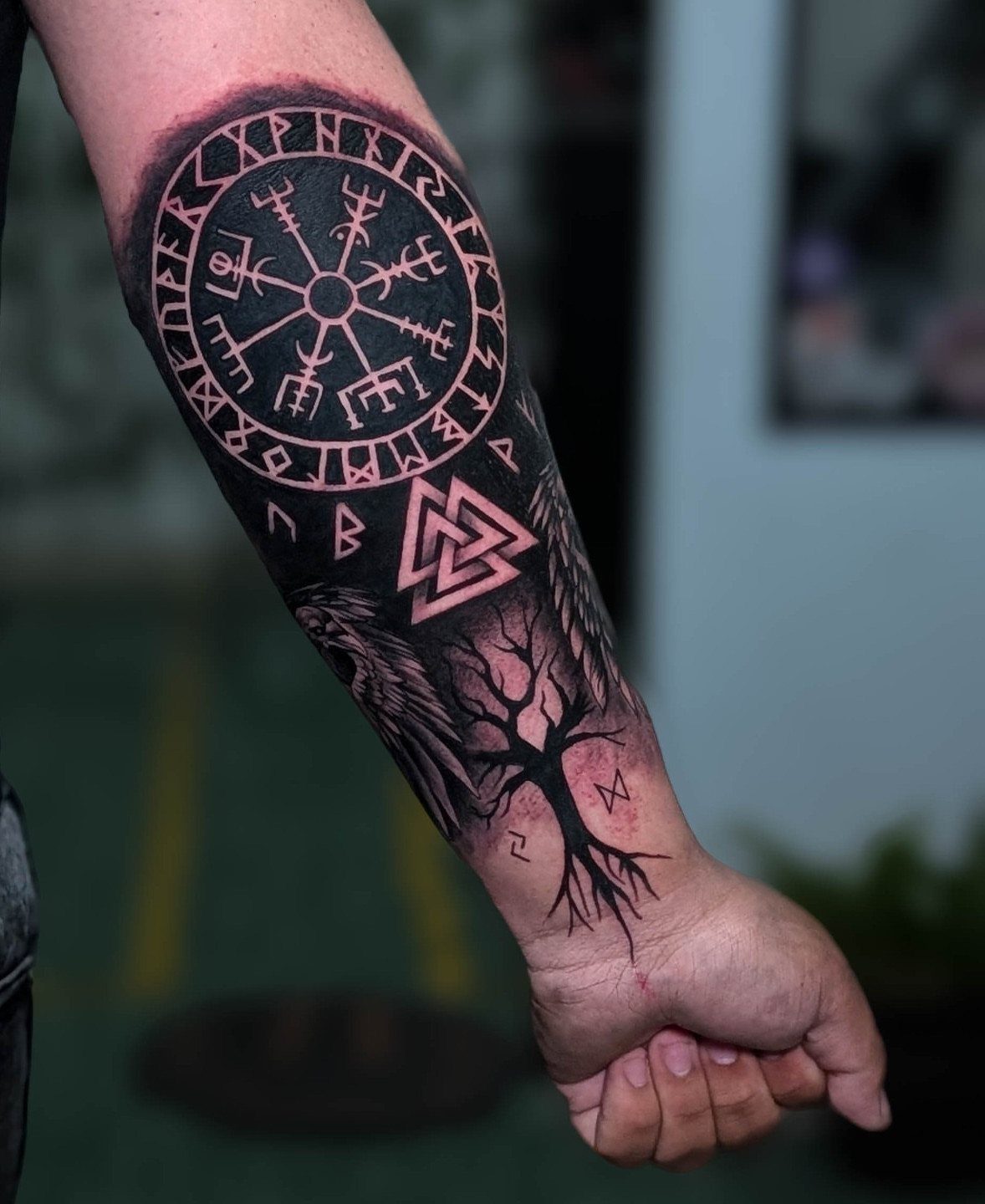 A person's forearm displaying a large black ink tattoo with circular runes and a Valknut symbol.