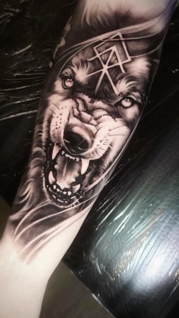 A detailed black and grey wolf tattoo with a Viking rune symbol on a forearm.