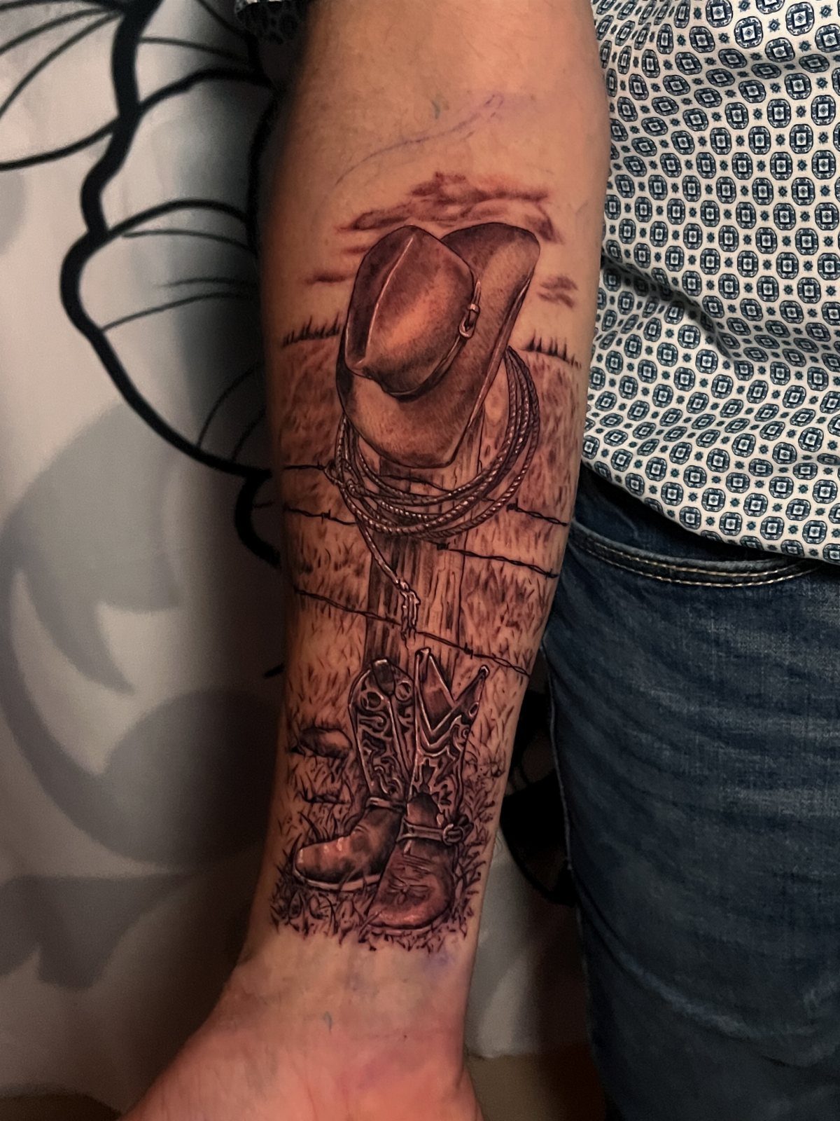 A black and grey forearm tattoo featuring a cowboy hat, lasso, and boots on a landscape background.