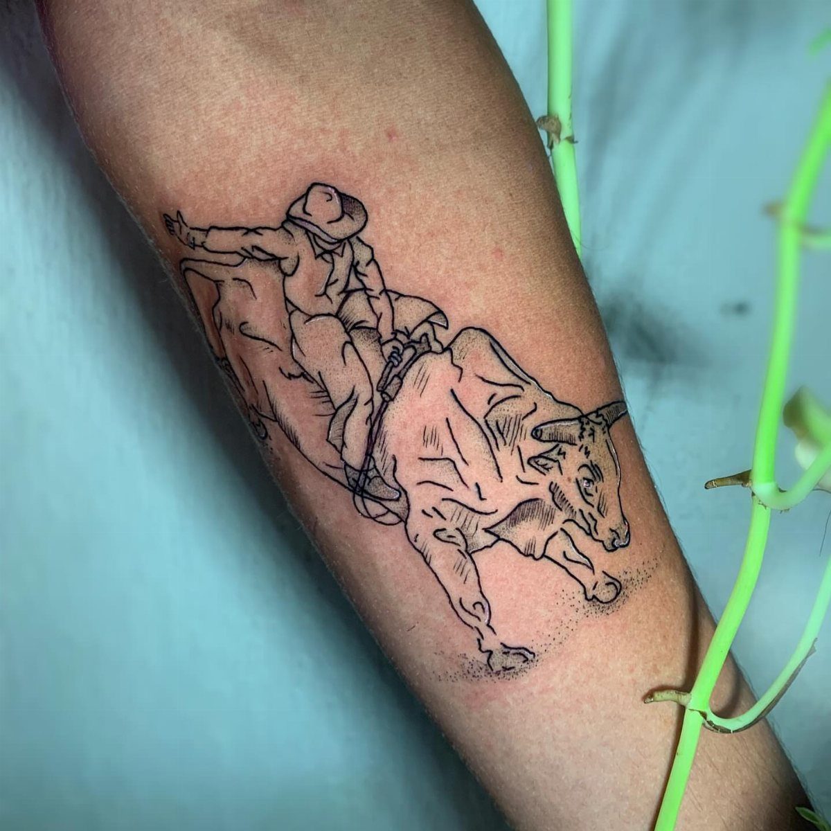 A black and grey tattoo of a cowboy riding a bucking bull on a forearm.