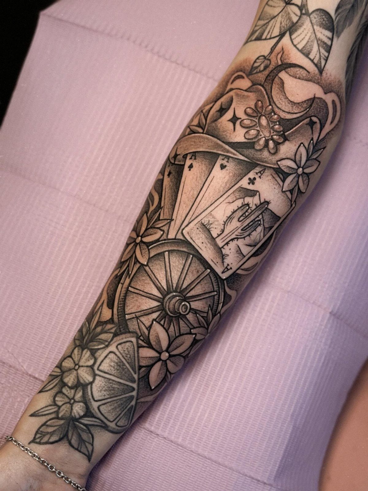A black and grey forearm tattoo featuring playing cards, a wagon wheel, and floral patterns.