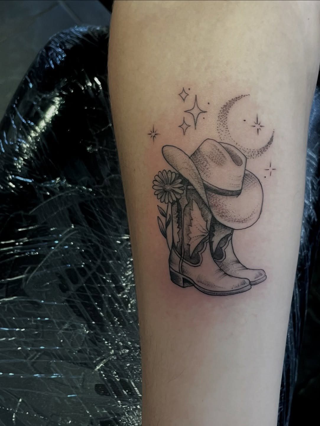A fine line tattoo of a cowboy hat, boot, flower, and crescent moon on skin.