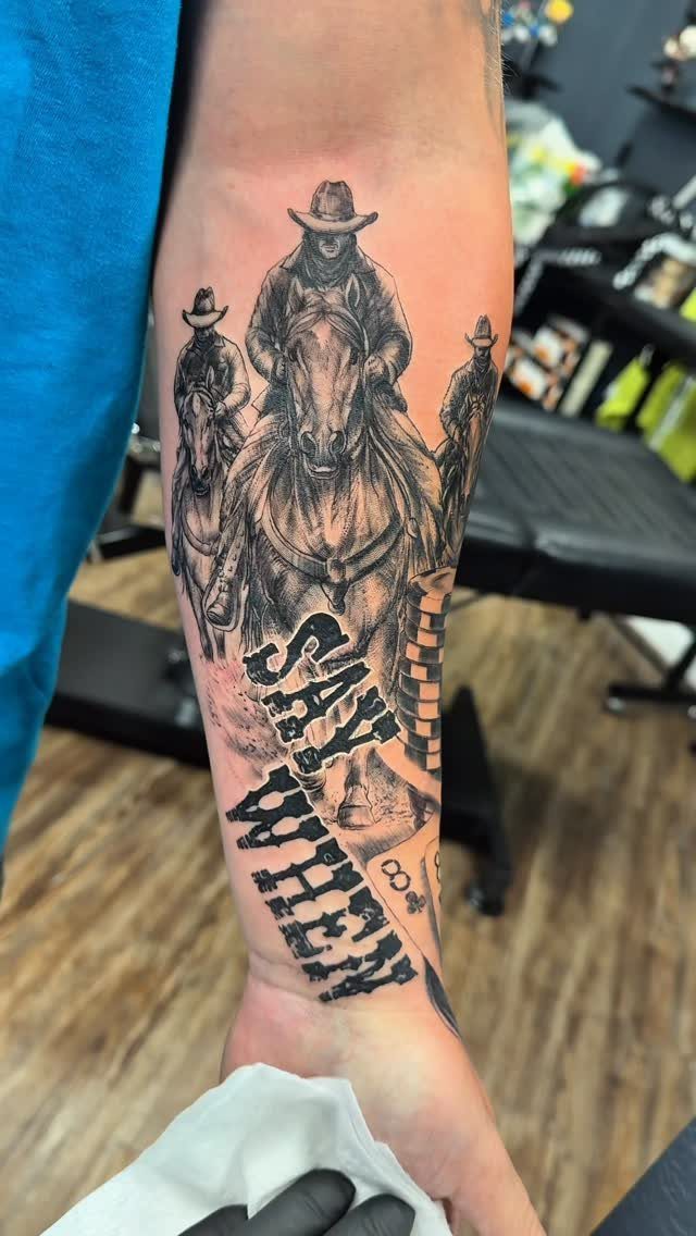 A black and grey fine line tattoo of cowboys riding horses on a person's forearm.