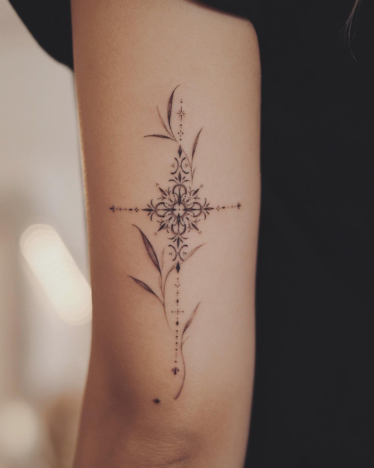 A fine line black ink tattoo of an ornamental cross with delicate vines on an arm.