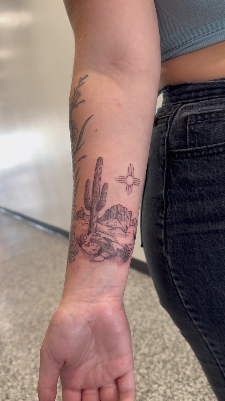 A fine line tattoo of a saguaro cactus and desert mountains on a person's forearm.