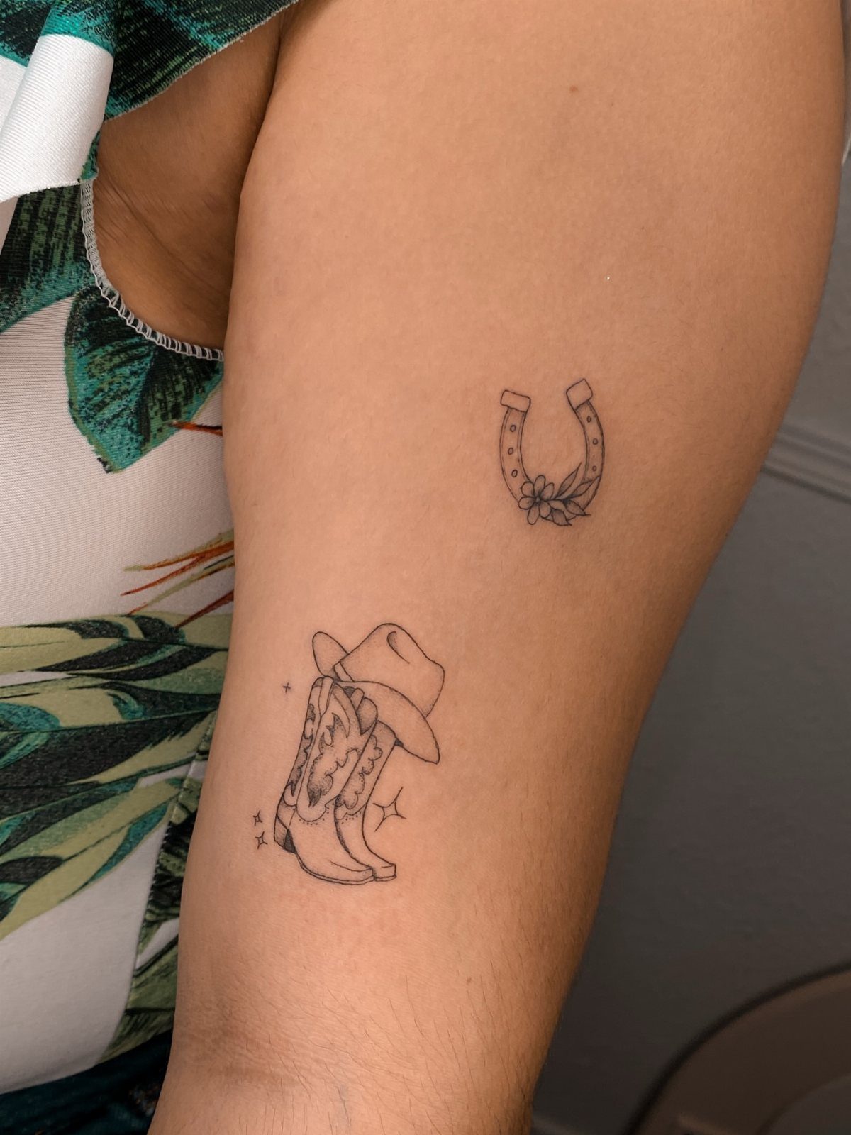 Two small black ink fine line tattoos on a person's arm, including a horseshoe and a cowboy boot.