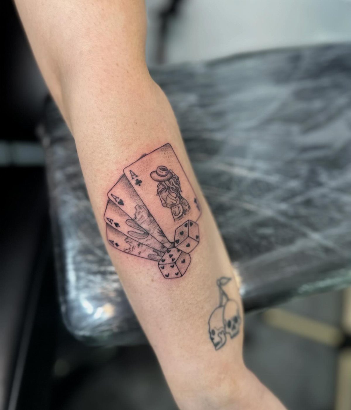 Fine line tattoo of playing cards, dice, and a cowboy hat on a person's forearm.