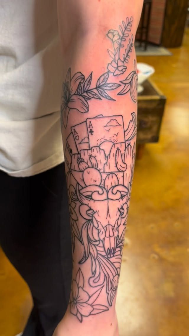 Black ink tattoo on a forearm featuring playing cards, mountains, and floral patterns.