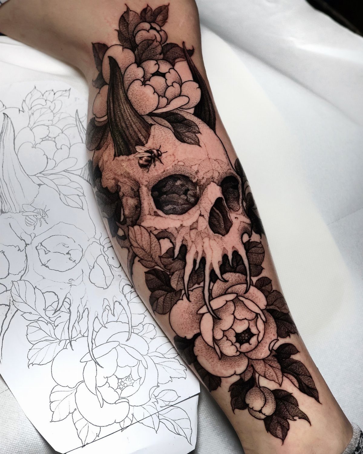 A black and grey forearm tattoo of a skull surrounded by large flowers and leaves.