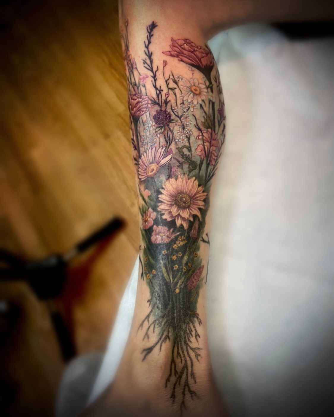 A detailed forearm tattoo of various pink and white wildflowers with long green stems and roots.